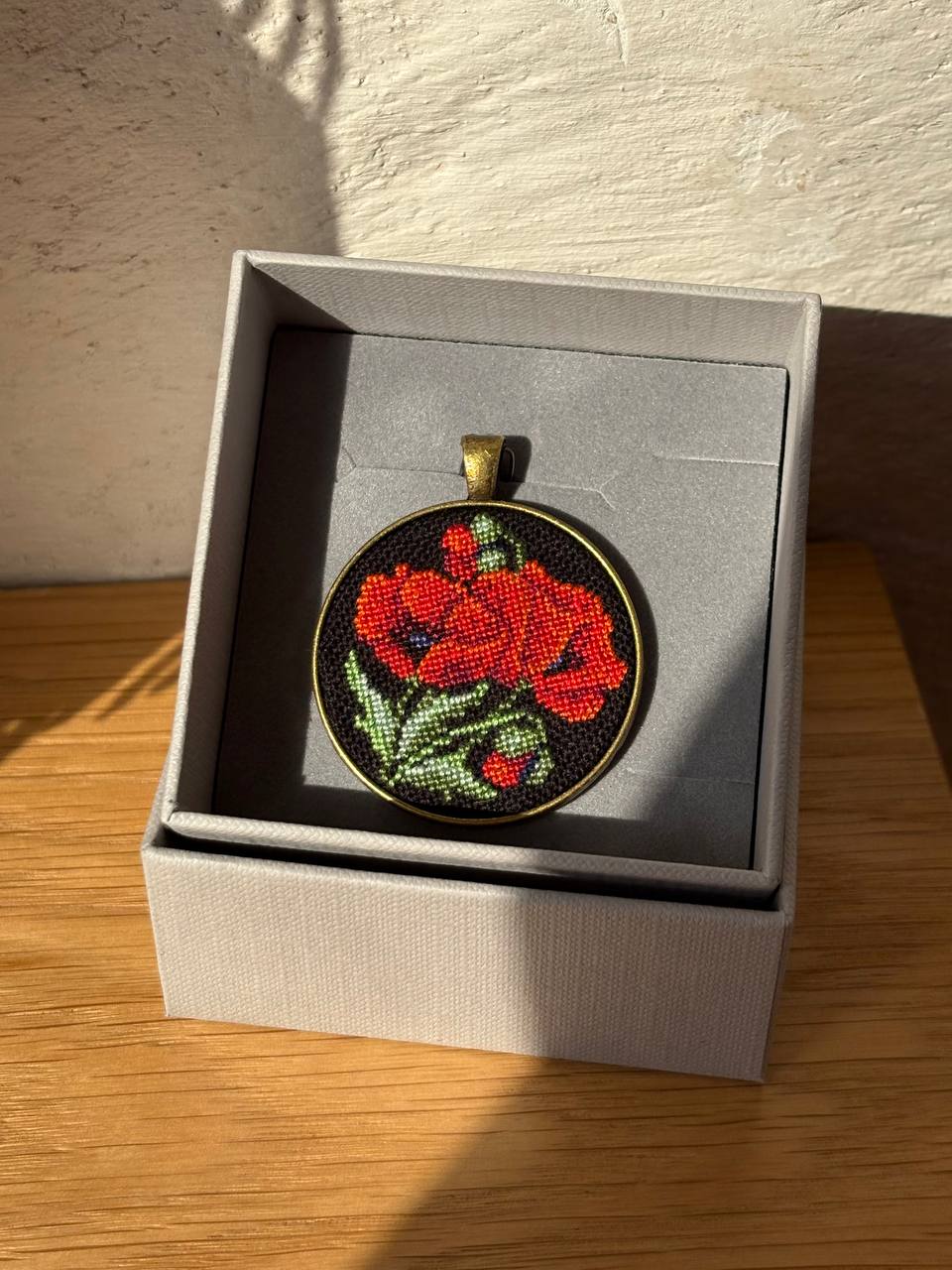 A round pendant with a red and green floral needlepoint design on a black background, displayed inside a gray presentation box on a wooden surface.
