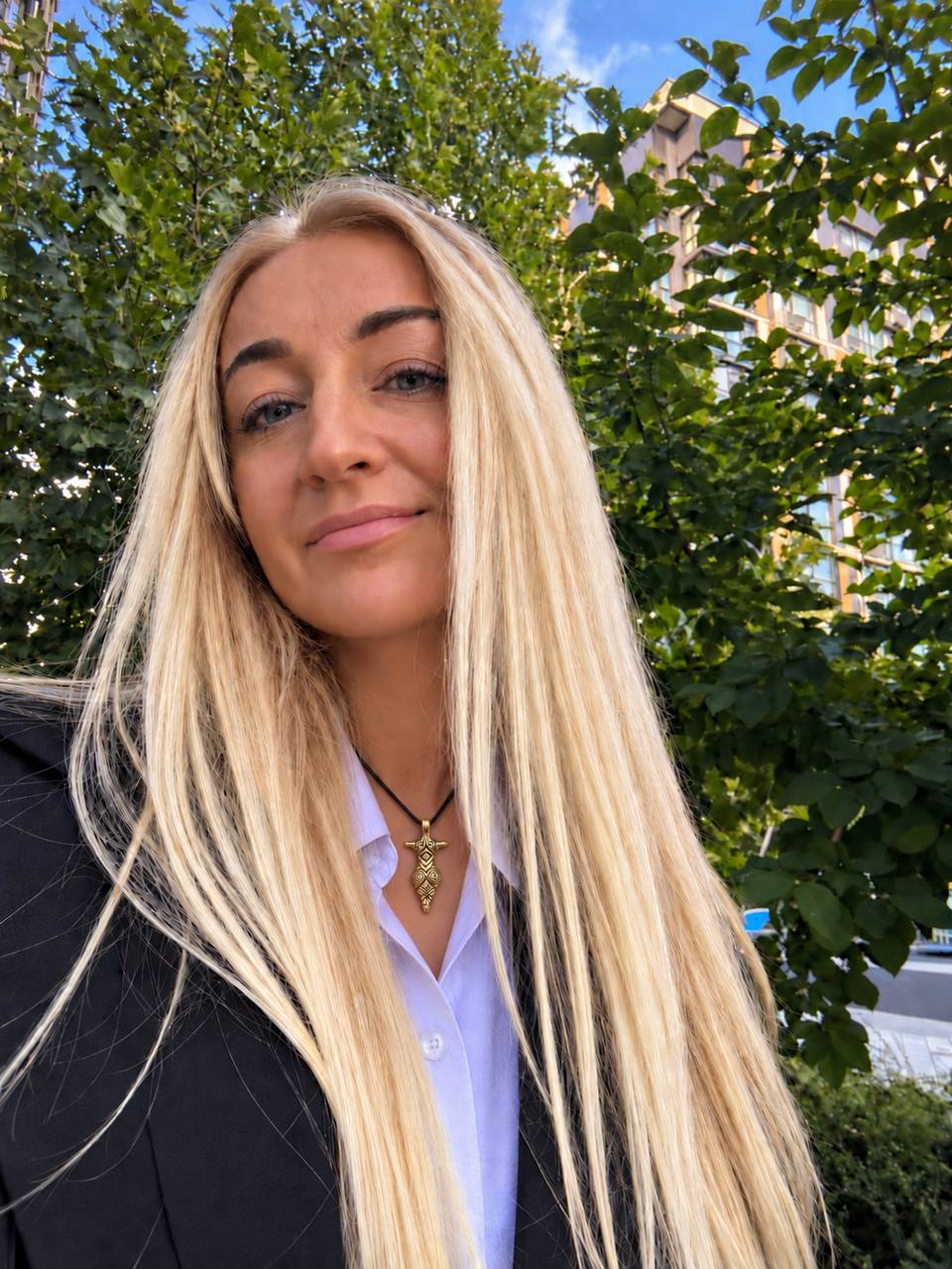 A woman with long, straight platinum blonde hair is shown in a close-up outdoor portrait. She is wearing a black blazer over a light blue collared shirt and a gold pendant necklace.