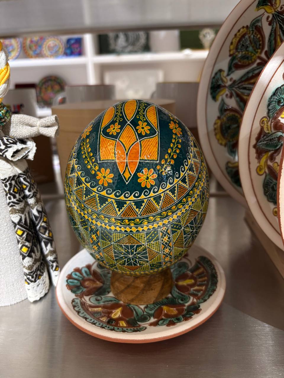 A hand-painted Ukrainian Easter egg with intricate geometric patterns in green, yellow, and orange, displayed on a matching ceramic saucer on a stainless steel counter.