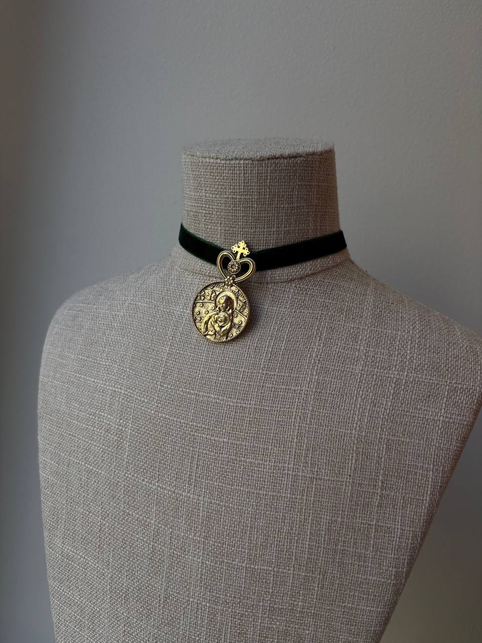 A black velvet choker necklace with a gold-tone circular pendant and small heart-shaped charm, displayed on a beige linen mannequin bust against a neutral gray background.