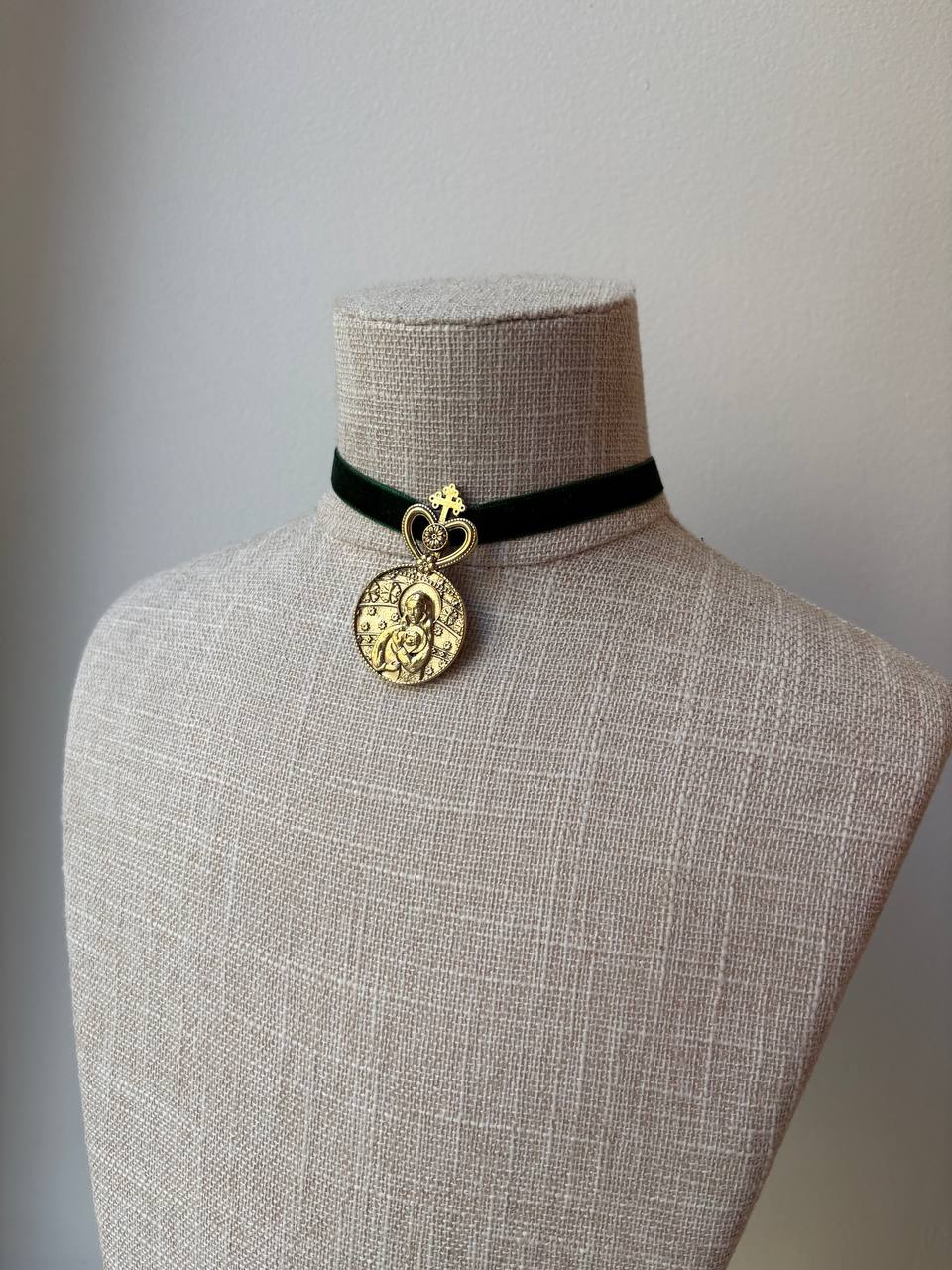 A black velvet choker necklace with a gold-tone metal heart-shaped connector and a round, textured gold pendant featuring a religious figure, displayed on a beige linen mannequin bust.