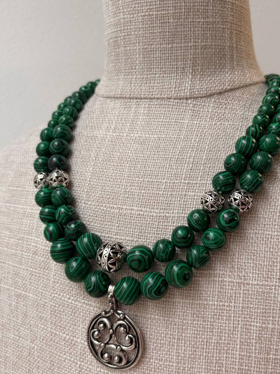 A double-strand green beaded necklace with malachite-like striped beads and silver filigree accents, displayed on a beige linen mannequin bust.