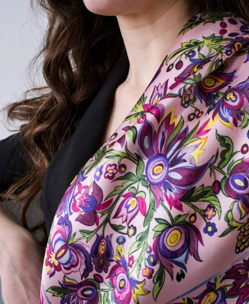 A close-up of a model wearing a light pink silk scarf with a vibrant, multicolored floral print in purple, green, yellow, and blue.