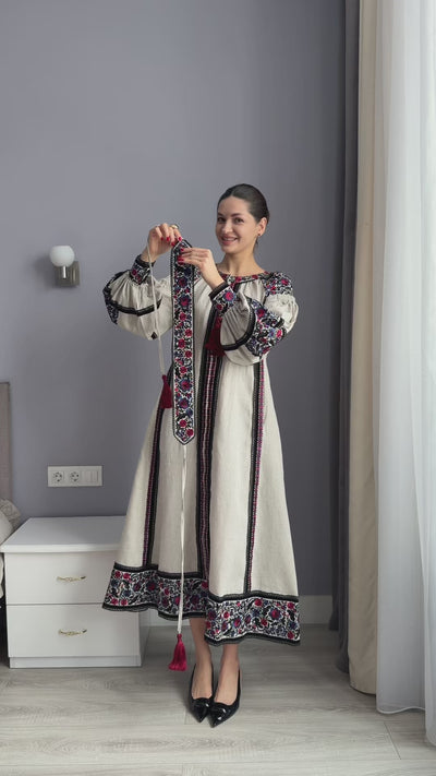 modeling a traditional long dress with cream-colored fabric, black and red embroidered details, and a red tassel tie, standing in a room with gray walls.