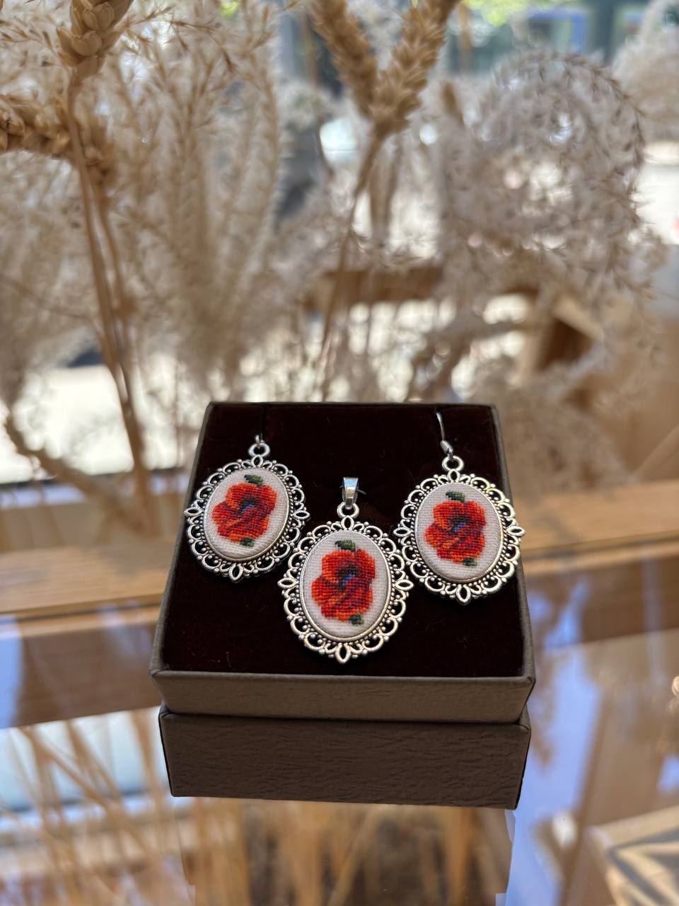 A set of embroidered earrings and pendant displayed in a box, featuring red floral patterns on a white background with a silver-colored frame.
