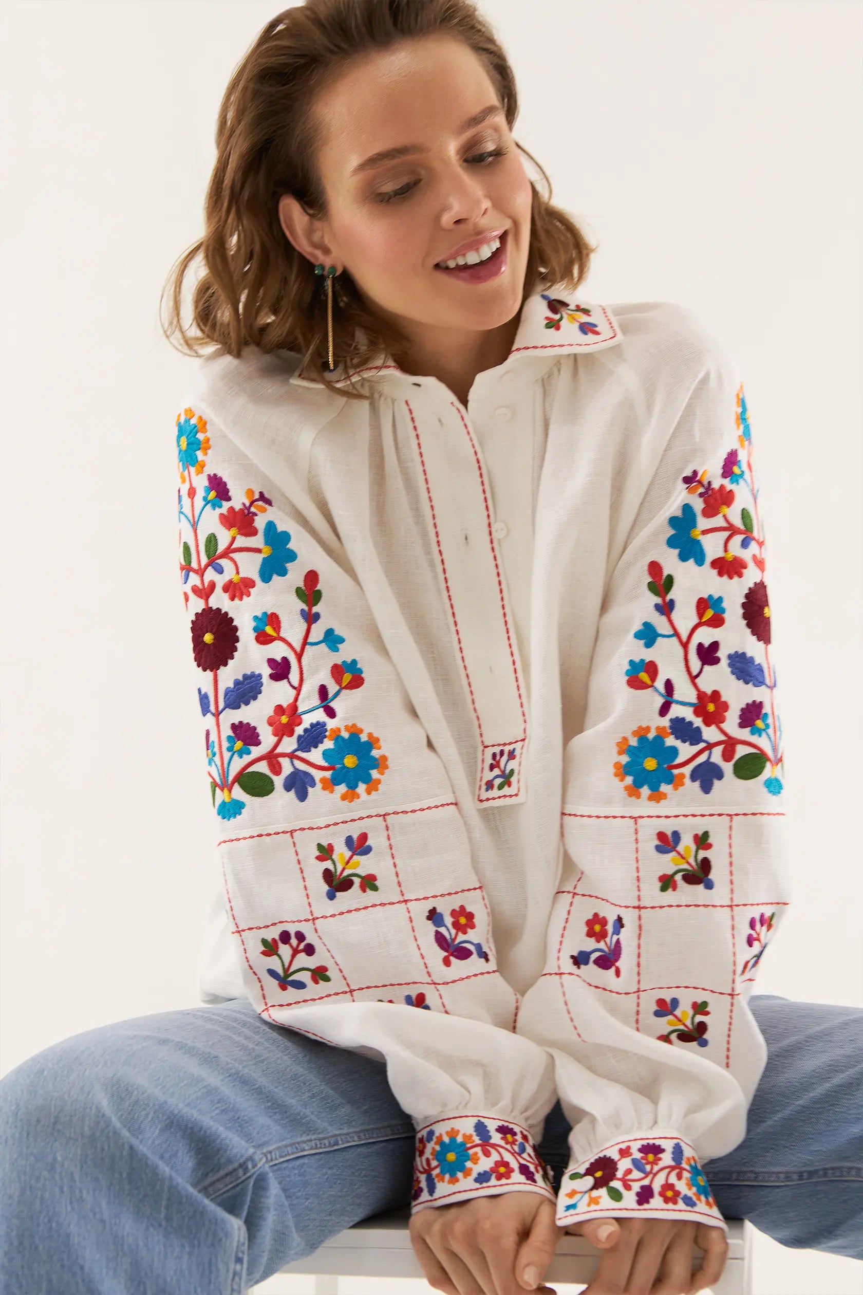 Woman wearing a white blouse with colorful floral embroidery sitting on a white surface.