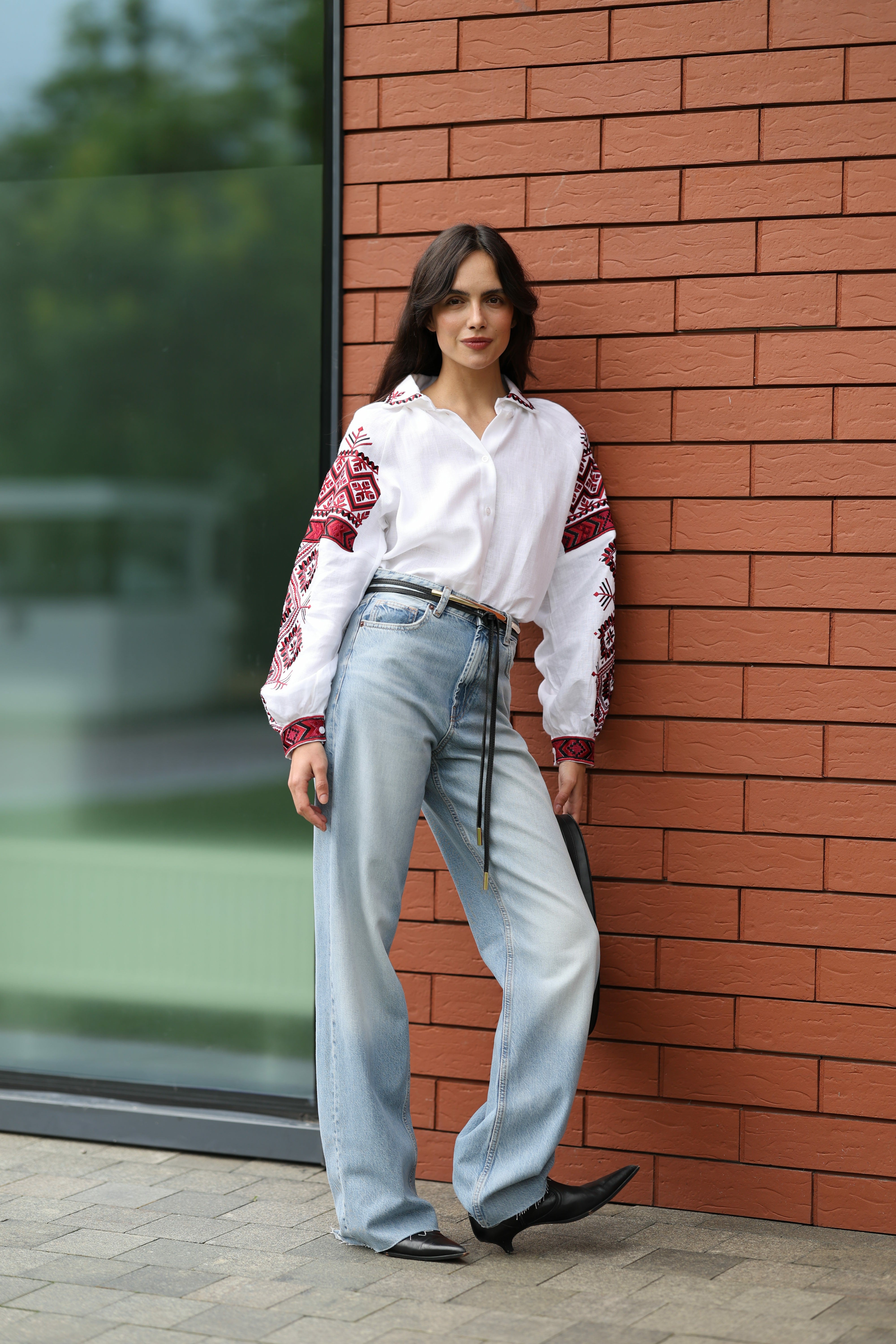 Woman in a white blouse with red patterns and blue jeans standing against a brick wall.