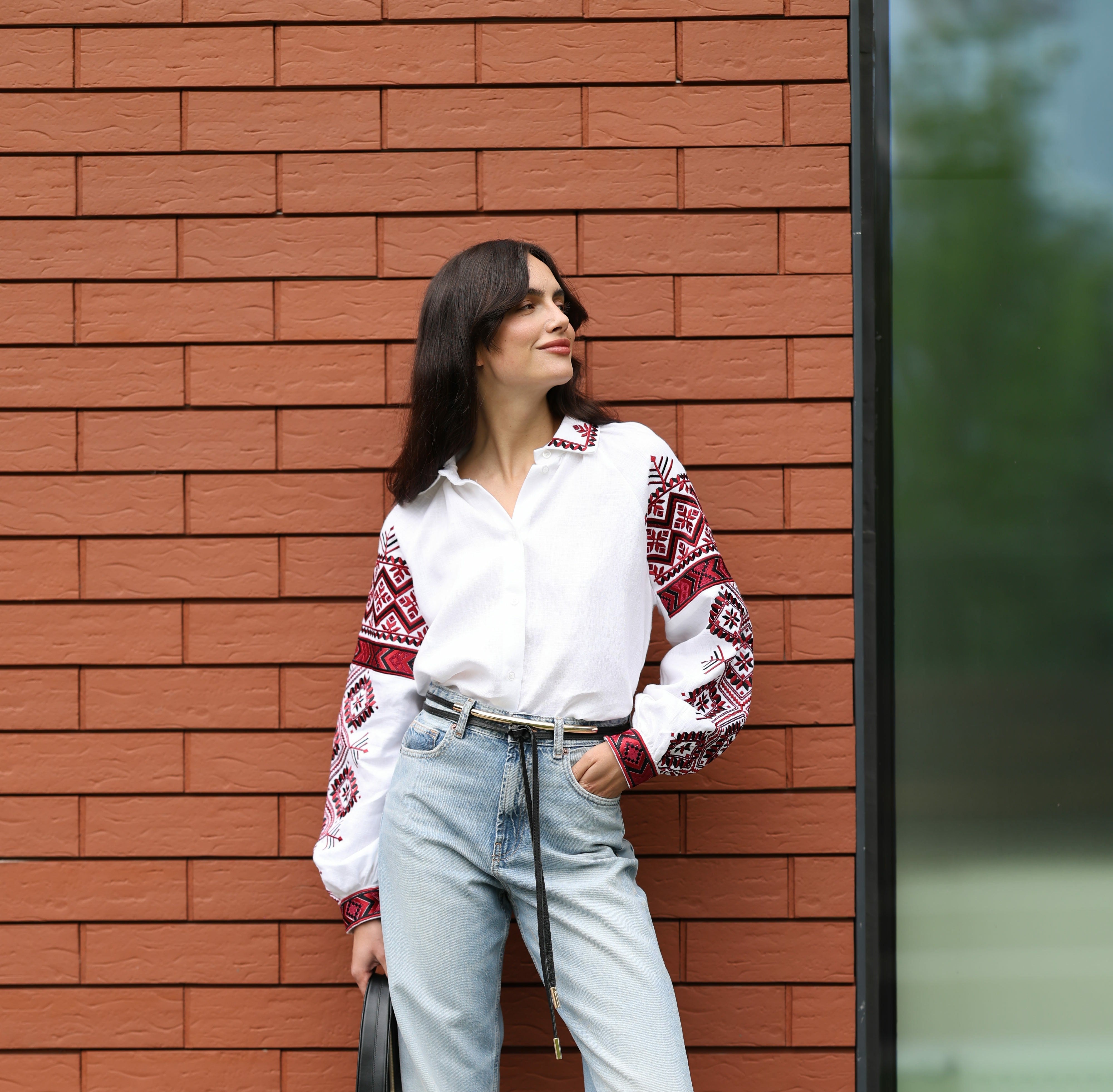 Woman in a white blouse with red and black patterns and light blue jeans standing against a brick wall.