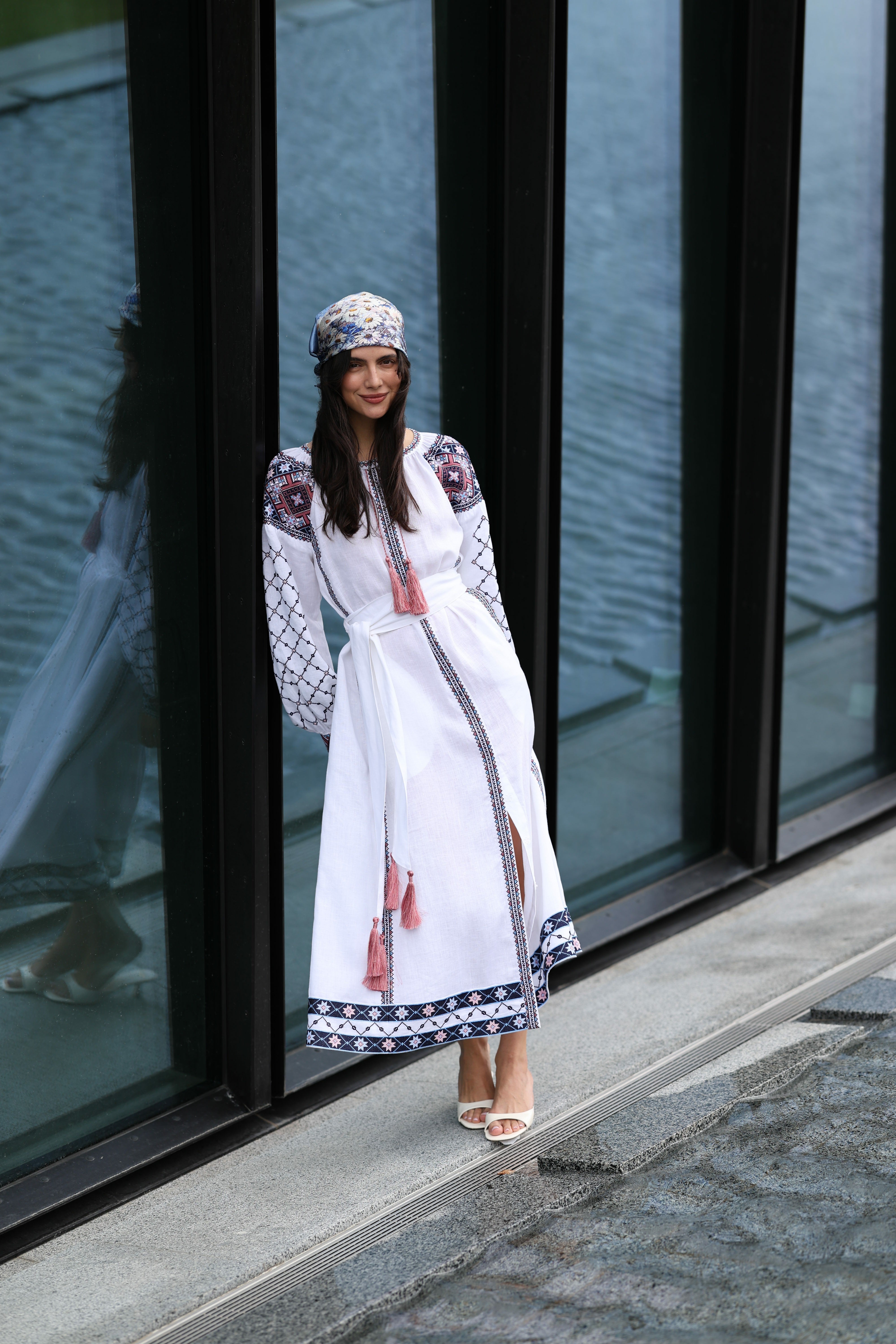Woman in a white dress with decorative patterns standing in front of a glass building.