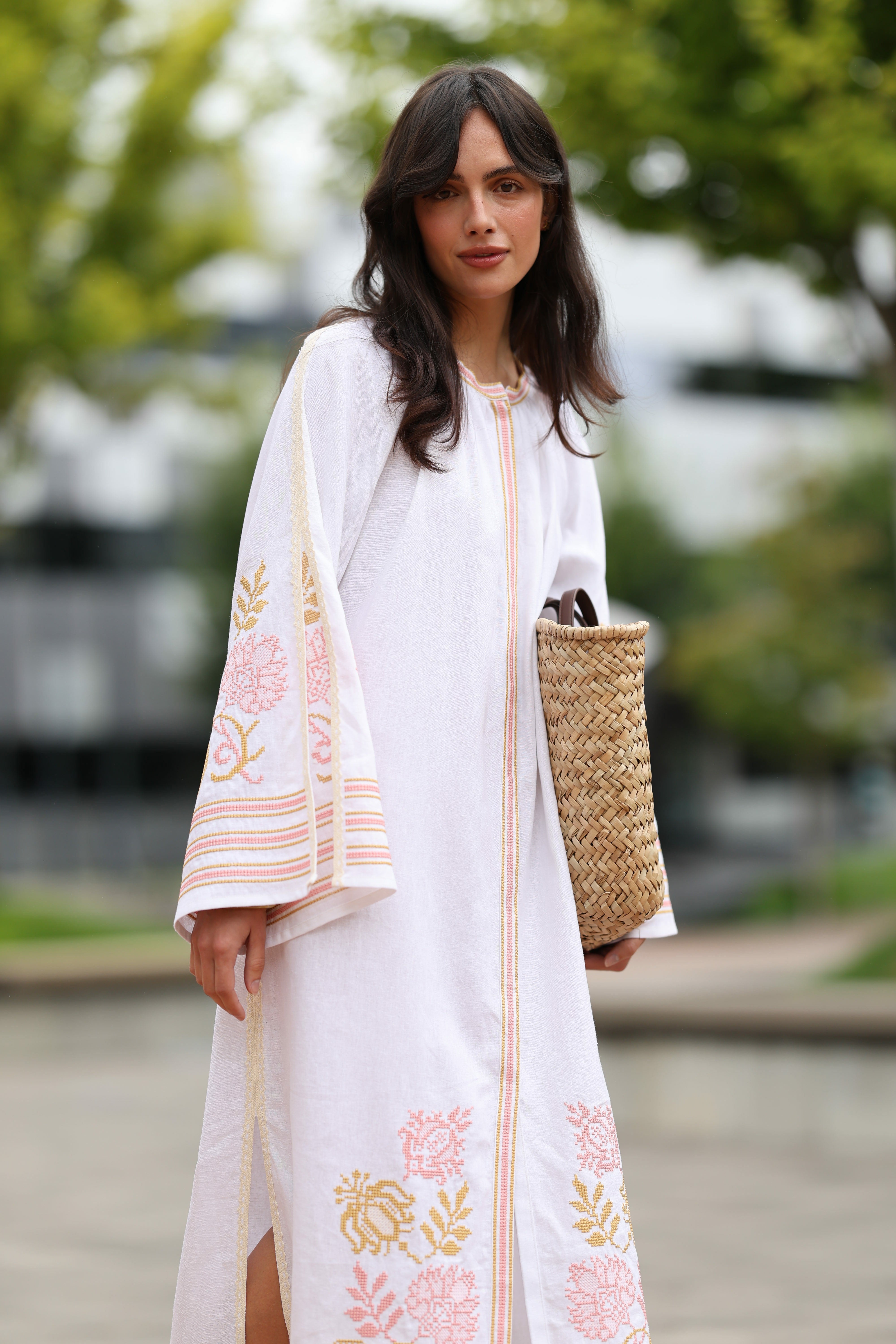 Woman wearing a white dress with floral embroidery, holding a woven bag outdoors.