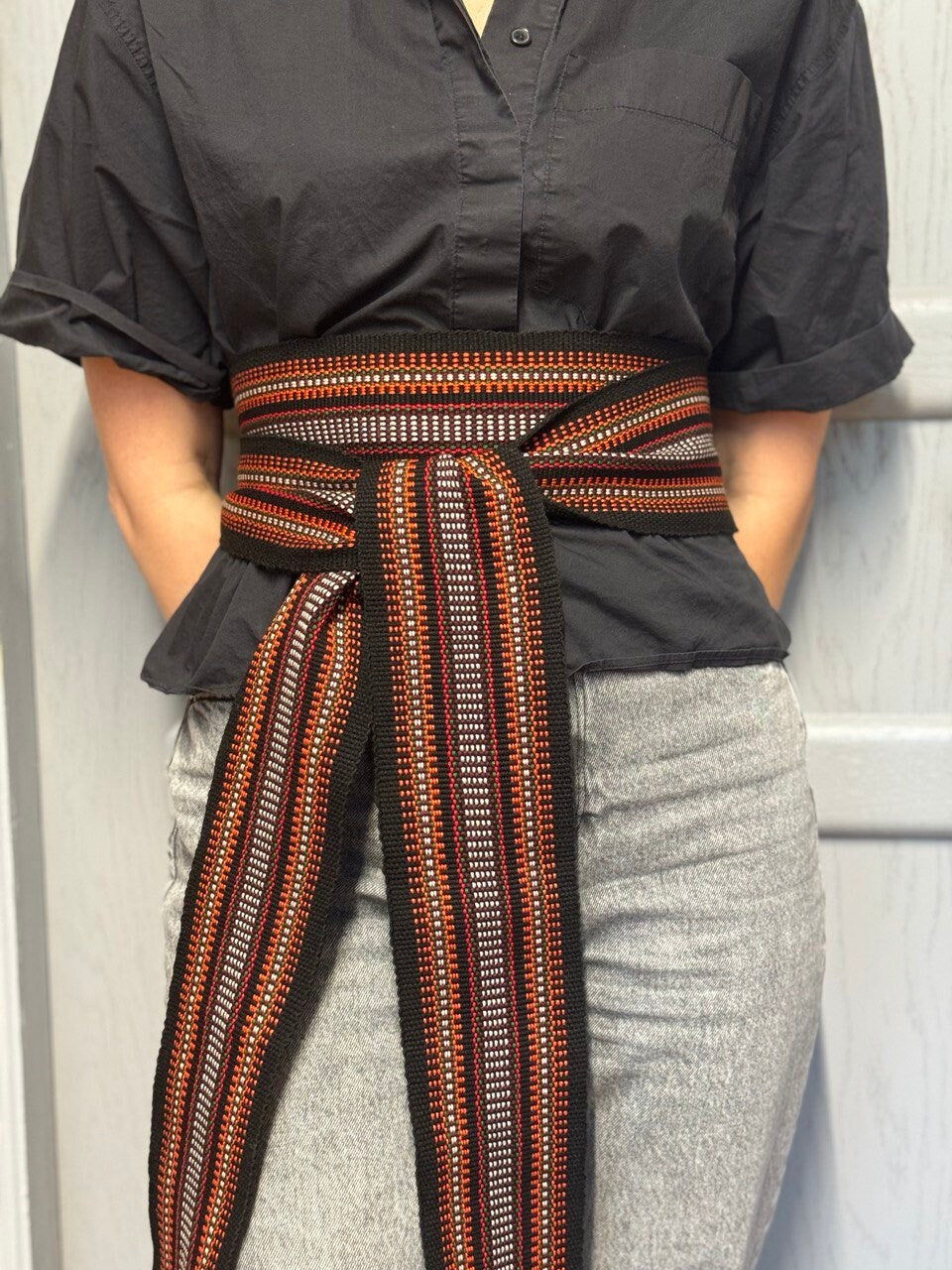 A person wearing a black shirt and gray pants with a woven belt with a geometric pattern tied around the waist.